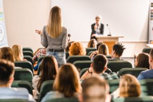 We see a female student standing up in a college classroom, asking her professor a question most likely pertaining to the servicenow platform.
