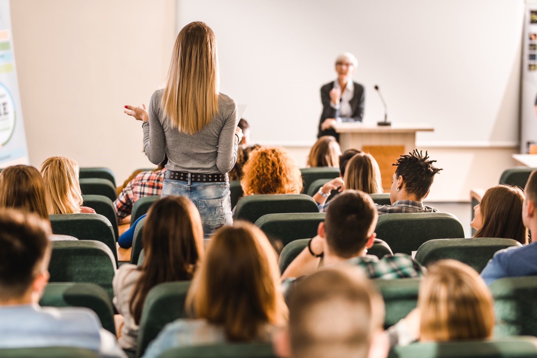 We see a female student standing up in a college classroom, asking her professor a question most likely pertaining to the servicenow platform.